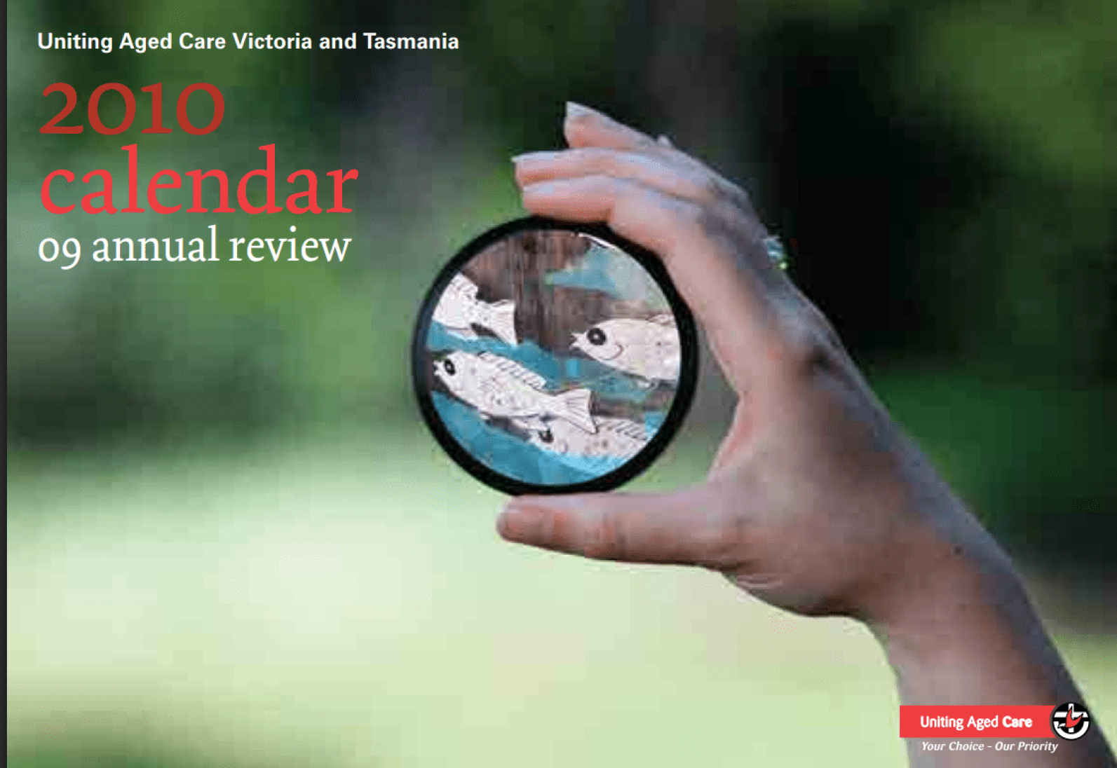 Cover of the Uniting Aged Care Victoria and Tasmania 2010 Calendar and 2009 Annual Review. A hand holds a round magnifying lens displaying an illustration of fish, set against a blurred green outdoor background. The title text appears in red and white, with the Uniting Aged Care logo and the tagline “Your Choice – Our Priority” in the bottom-right corner.