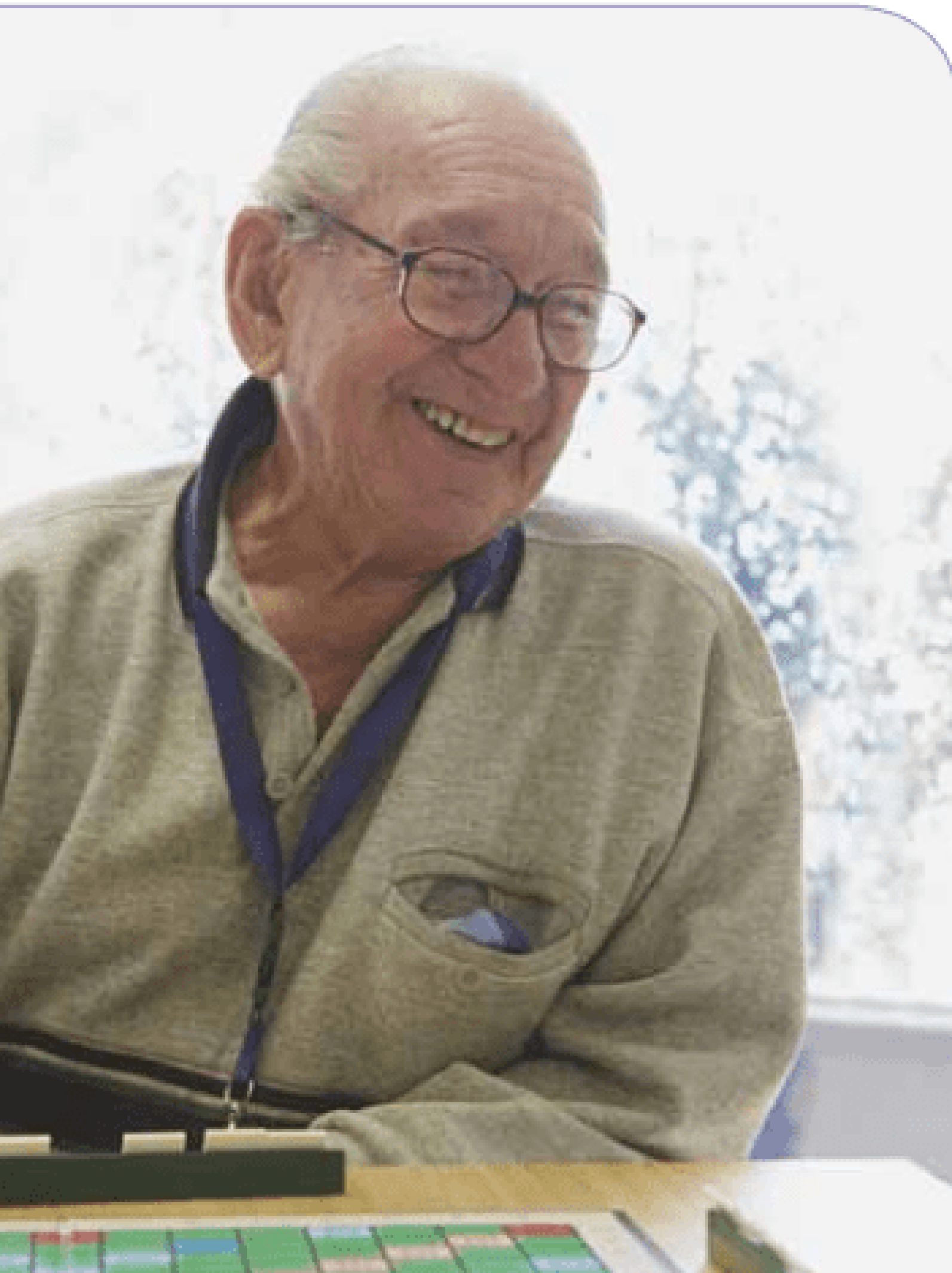 An older adult seated at a table playing a board game with letter tiles. Wearing glasses and a lanyard, the person is smiling while looking toward the game board. Natural light streams through a nearby window, creating a bright and welcoming setting that encourages social interaction and recreational activities.