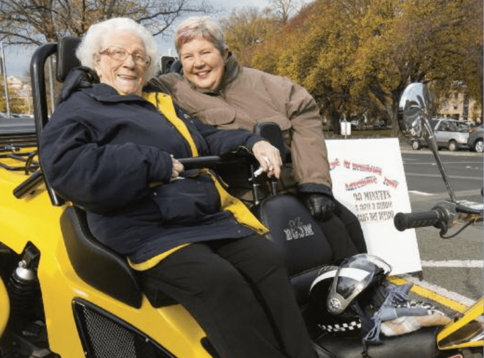 An older adult seated on a bright yellow three-wheeled motorcycle-style vehicle, accompanied by another adult standing closely beside them. The pair are outdoors in a parking area with trees and vehicles in the background. A motorcycle helmet rests on the vehicle, highlighting participation in a community outing and shared recreational experience.