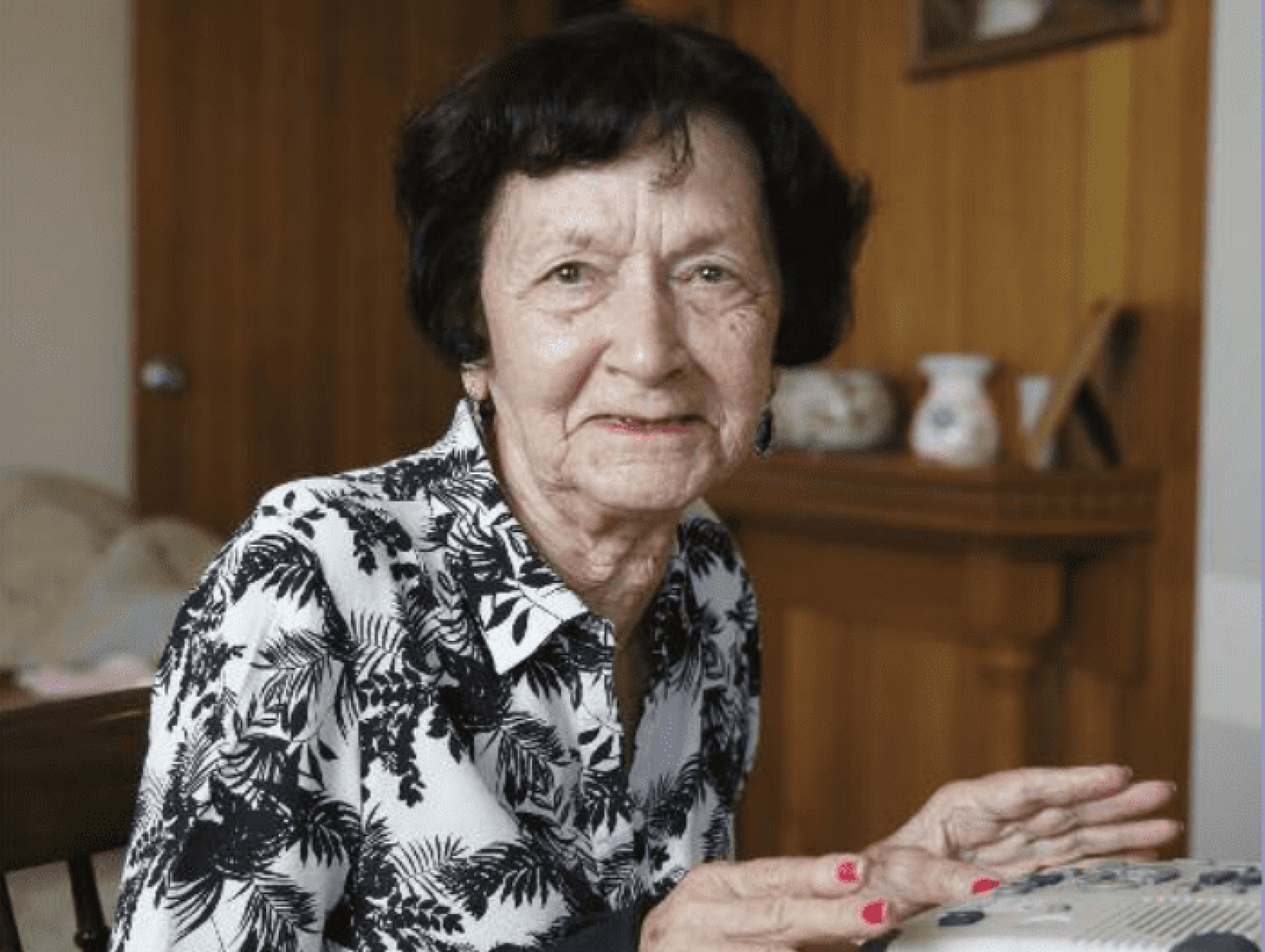 An older adult seated indoors at a table, looking toward the camera. The person is wearing a black-and-white patterned blouse, with hands resting near an electronic keyboard or activity device. Timber furnishings and decorative items in the background create a warm and homely living environment.