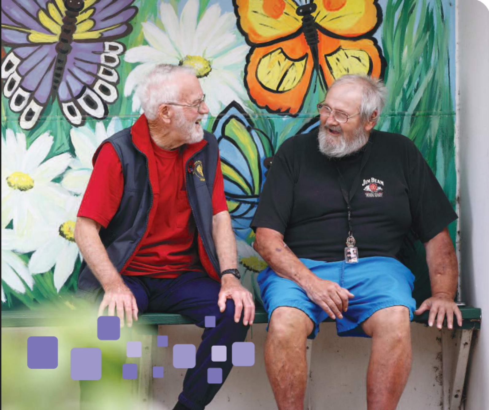 Two older adults seated side by side on a bench in front of a colourful mural featuring large butterflies and flowers. The pair are engaged in conversation in an outdoor community setting, with the vibrant artwork creating a cheerful and welcoming backdrop.