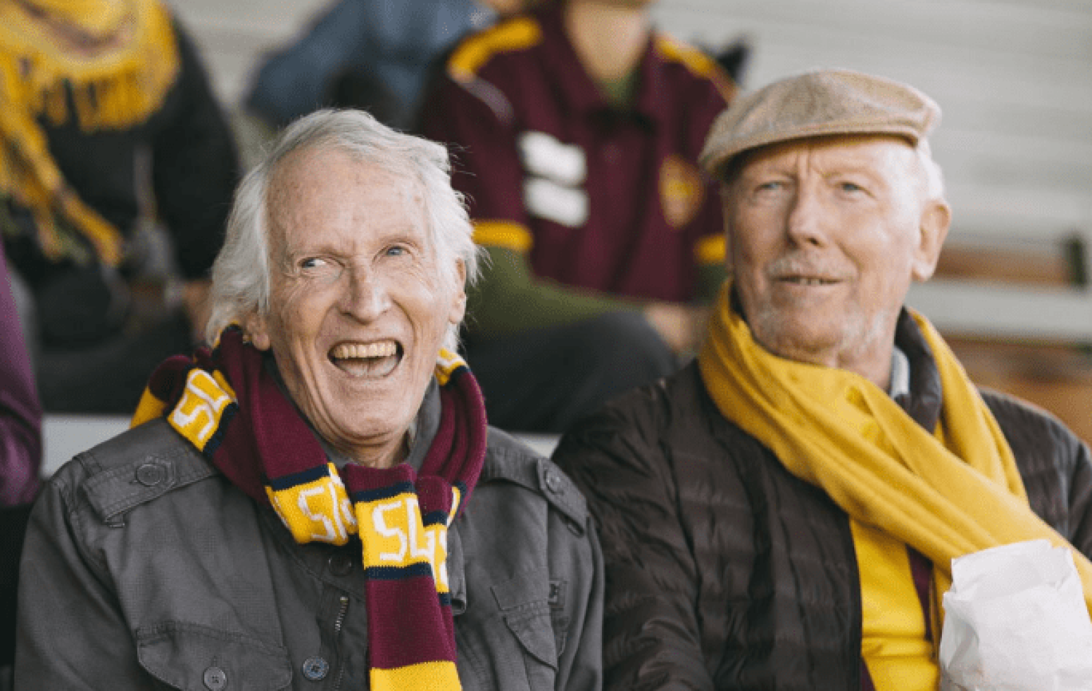 Two older adults seated in outdoor stadium-style seating, wearing team scarves and jackets while watching a sporting event. Other spectators in matching team colours are visible in the background, highlighting community participation, recreation and enjoying live sport together.