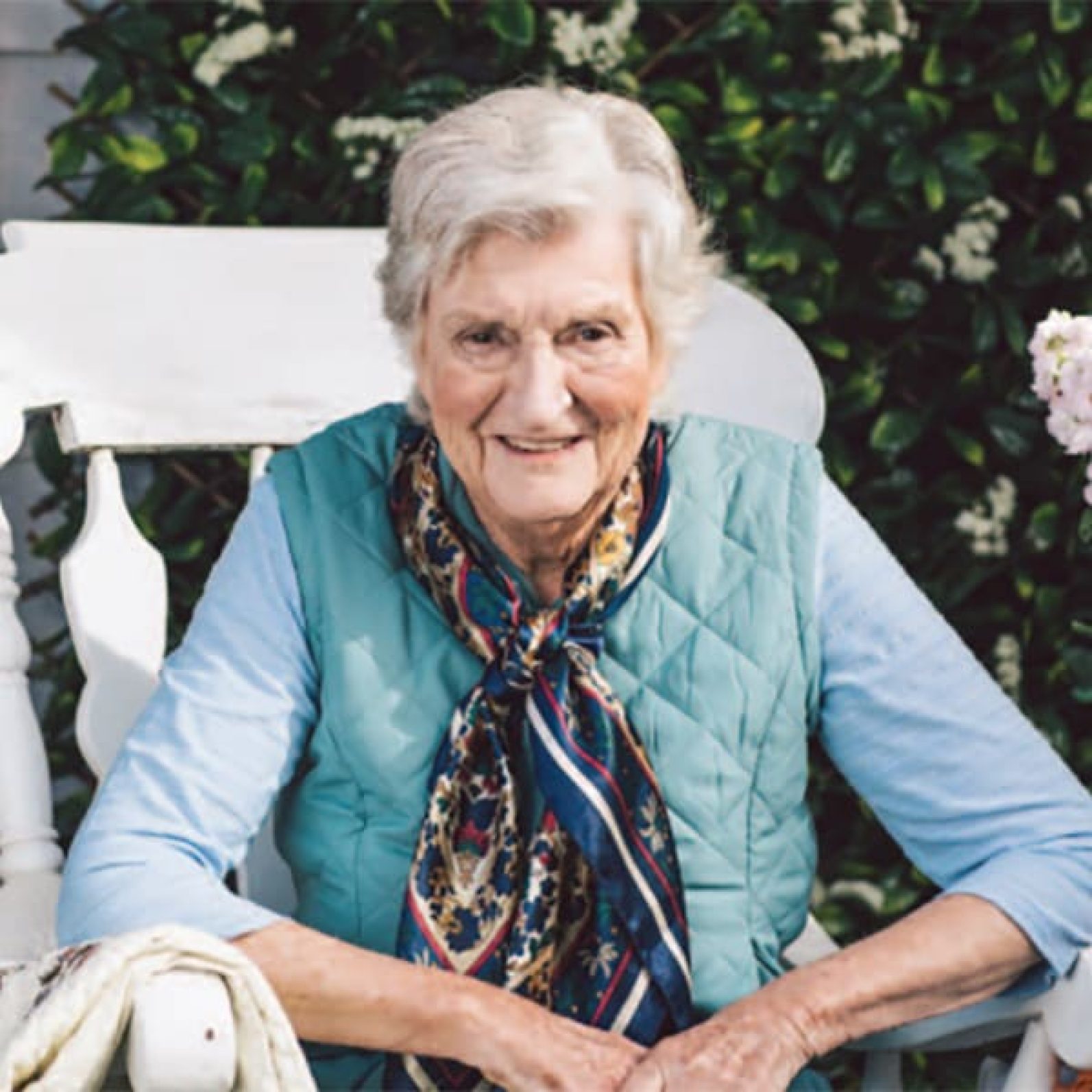 An older adult seated outdoors on a white garden bench, wearing a light blue quilted vest and patterned scarf. Flowering greenery and foliage form a natural backdrop, creating a peaceful garden setting that highlights relaxation and enjoyment of the outdoors.