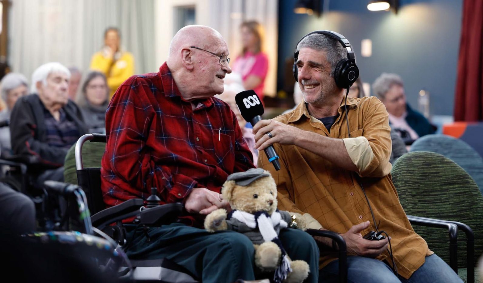 A broadcaster wearing a headphone is interviewing a resident with a microphone. The resident is holding onto a teddy bear.