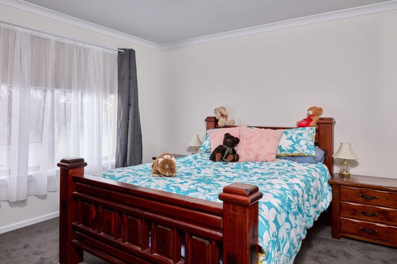 A bedroom is photographed, there is a bed in the middle of the room and windows on the side.