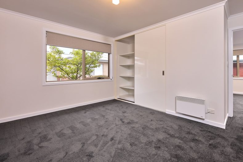 A bedroom is photographed, featuring a medium sized window and built-in wardrobe.