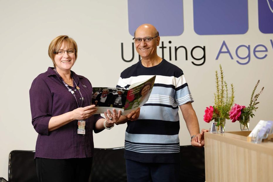 A staff member and a customer are pictured at the entrance of the community hub. The Uniting AgeWell logo is visible on the wall in the background.