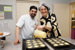 A chef and a broadcaster are smiling at the camera in the kitchen area as they are preparing food.