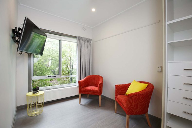 Premium suite sitting area, featuring two red chairs, a television attached to the wall next to the window where sunlight shines into the room
