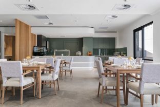 The dining area features wooden tables with four seats foor each of them. On the tables are cutleries and there is a kitchen area in the background.