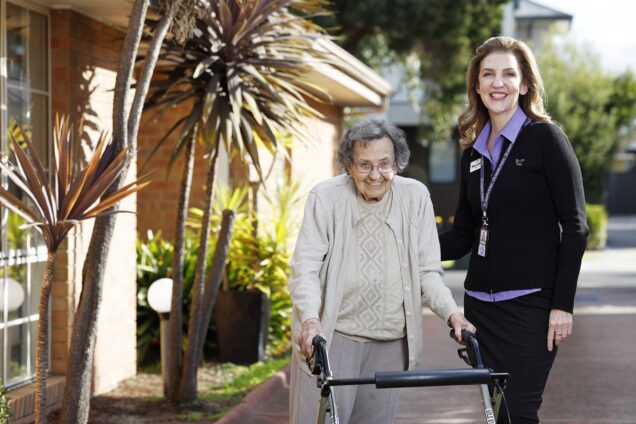 An older adult using a four-wheeled walker stands beside a staff member on a sunny pathway outside a residential aged care community. Surrounded by landscaped gardens and tropical plants, the pair are positioned near the building entrance, highlighting support, mobility and independence in a welcoming outdoor environment.
