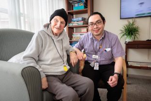A resident and a staff member aare sitting at the lounge, looking at the camera