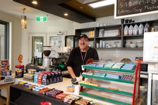 A staff member is smiling at the camera from the counter. There are chocolate and sweets for sale in front of the staff member. Next to them is a coffee machine.