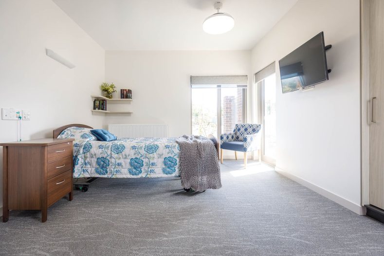 Well-lit room with bed, TV, bedside drawerer, sofa chair and wall shelf.