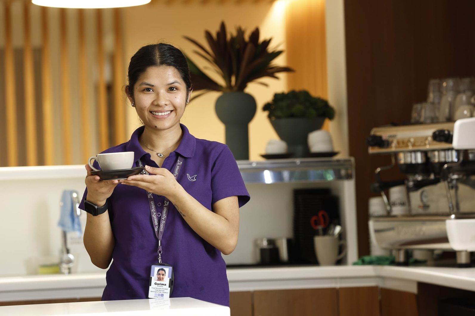 A smiling staff member holds up a cup, with a coffee machine pictured in the background