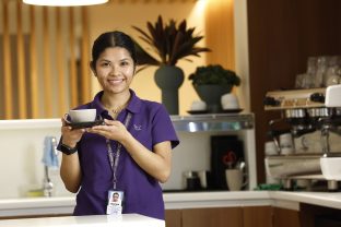 A smiling staff member holds up a cup, with a coffee machine pictured in the background