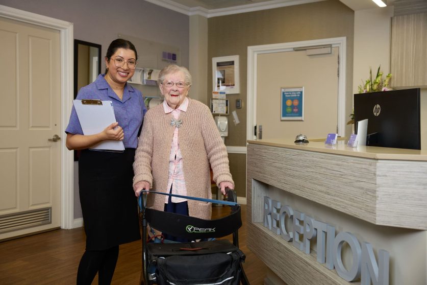 The image captures a staff member and a resident standing at the reception of Manor Lakes Community.