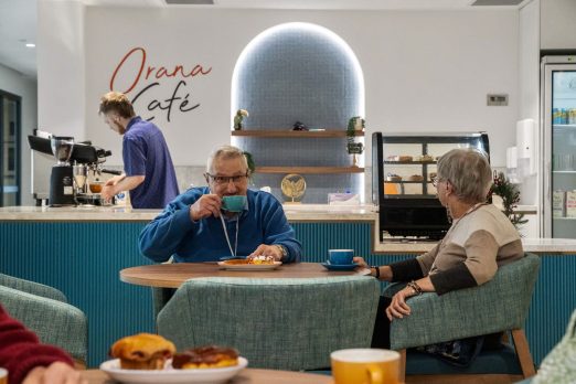 A few people are sitting in the cafe to enjoy their food and drinks. Behind them is a kitchen area where a staff member is seen making coffee