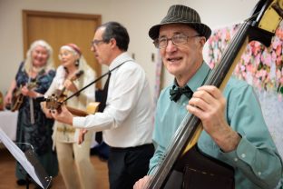 A person in sky-blue shirt with a black hat is holding their cello and smiles at the camera, people in the background are playing music.