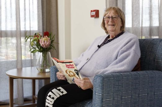 A resident is sitting next to the window with a magazine in hand, smiling at the camera