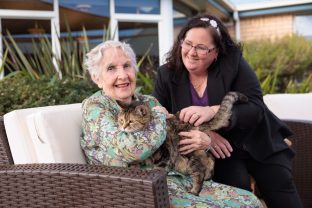A resident smiles at the camera while holding onto her cat, another person looks at the resident smiling.