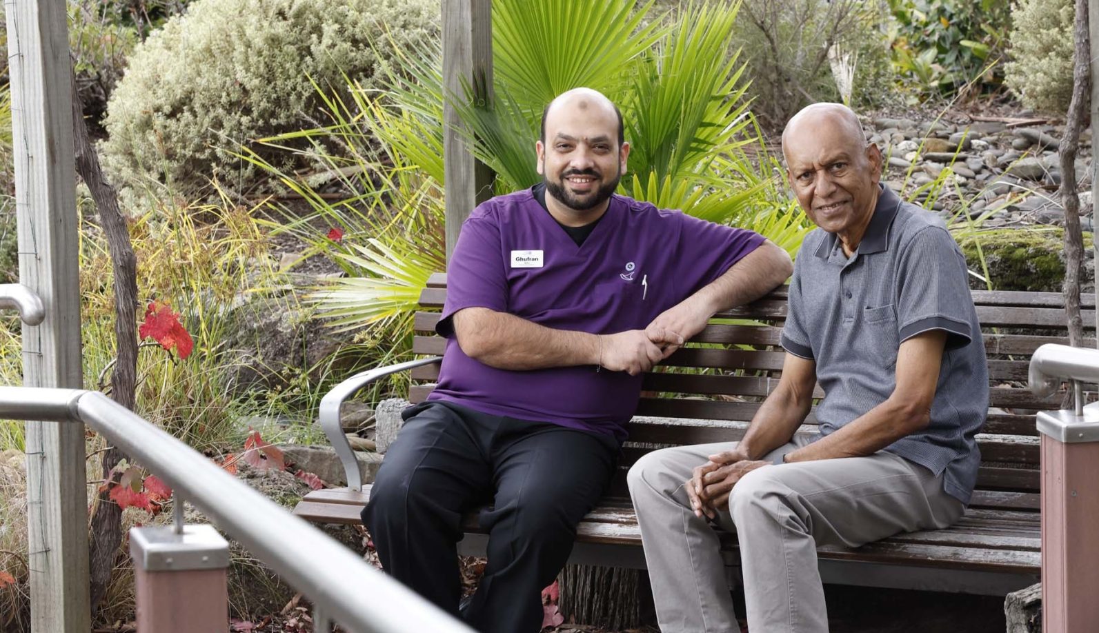 A resident and a staff member are siitting on a bench in the garden area, smiling at the camera