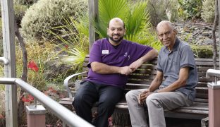 A resident and a staff member are siitting on a bench in the garden area, smiling at the camera