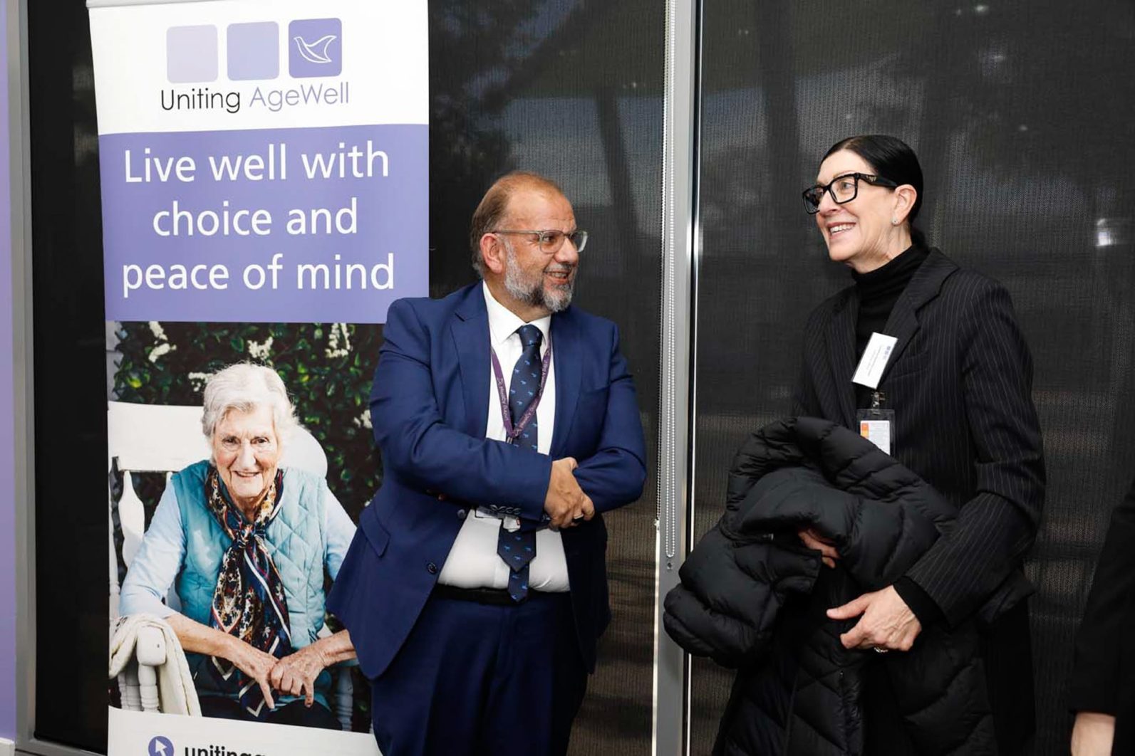 Two people are photographed chatting, there is a Uniting AgeWell banner on the side.