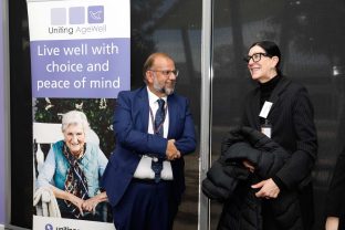 Two people are photographed chatting, there is a Uniting AgeWell banner on the side.