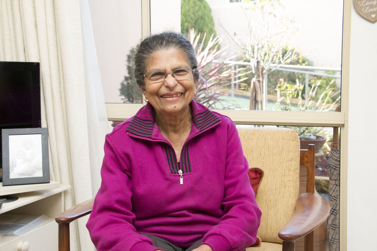 A person wearing glasses and a bright magenta jumper sits in a wooden armchair indoors. Large windows behind the chair overlook a garden with plants and shrubs, while a television, framed photograph and light-coloured curtains are visible in the room. Natural light fills the comfortable living space.