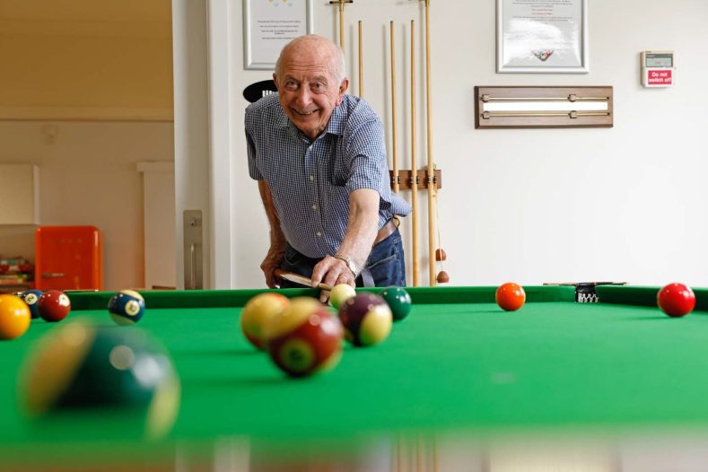 A man is playing billards, looking at the camera which is taking the photo from the table perspective.