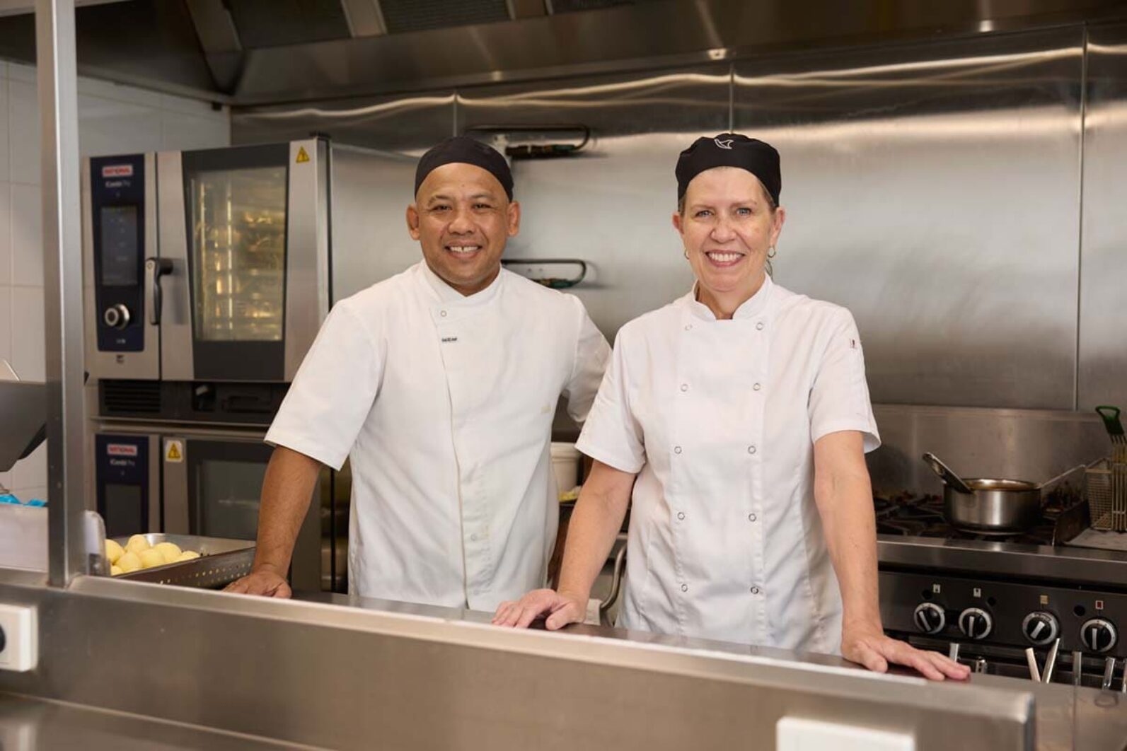 Two chefs in the kitchen smiling at the camera.
