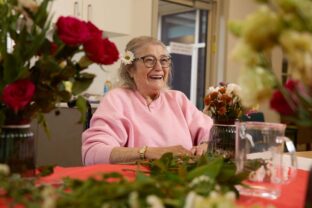 A resident smiling during a flower arranging workshop, with a flower tucked beside her hair.