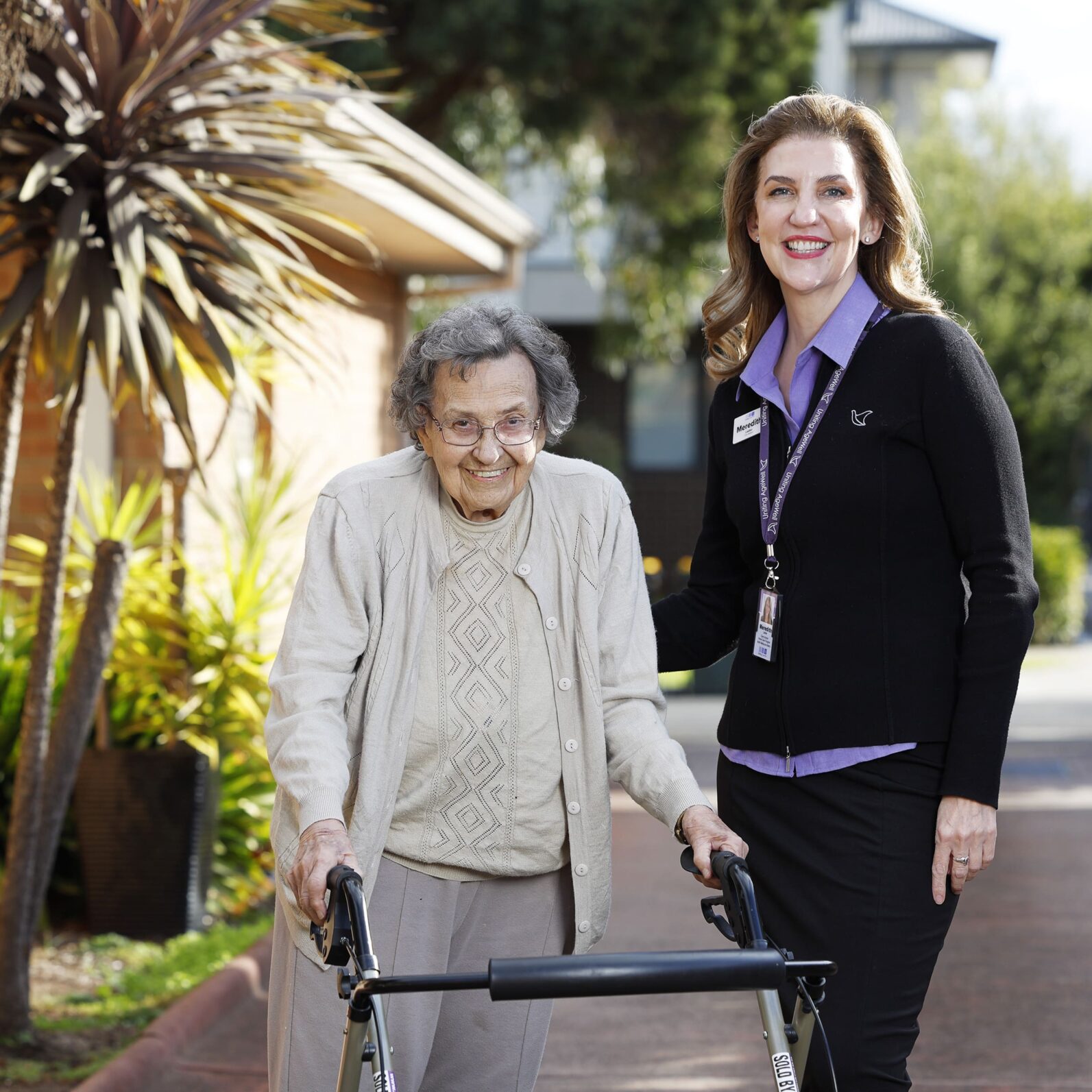 An older adult and a staff member stand together outdoors on a landscaped pathway. The older adult uses a four-wheeled walker, while the staff member stands alongside with a hand gently resting on the walker. Trees, garden plants and a residential-style building create a bright and welcoming setting that highlights independence, mobility and personalised support.
