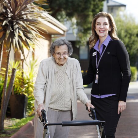 An older adult and a staff member stand together outdoors on a landscaped pathway. The older adult uses a four-wheeled walker, while the staff member stands alongside with a hand gently resting on the walker. Trees, garden plants and a residential-style building create a bright and welcoming setting that highlights independence, mobility and personalised support.