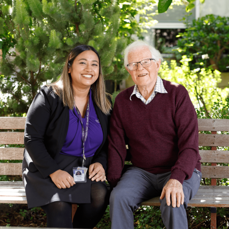 A staff member wearing a purple top and lanyard sits beside an older adult on a wooden garden bench surrounded by lush greenery. Both are seated outdoors in a landscaped courtyard, facing the camera in a welcoming setting that highlights companionship, connection and personalised support.