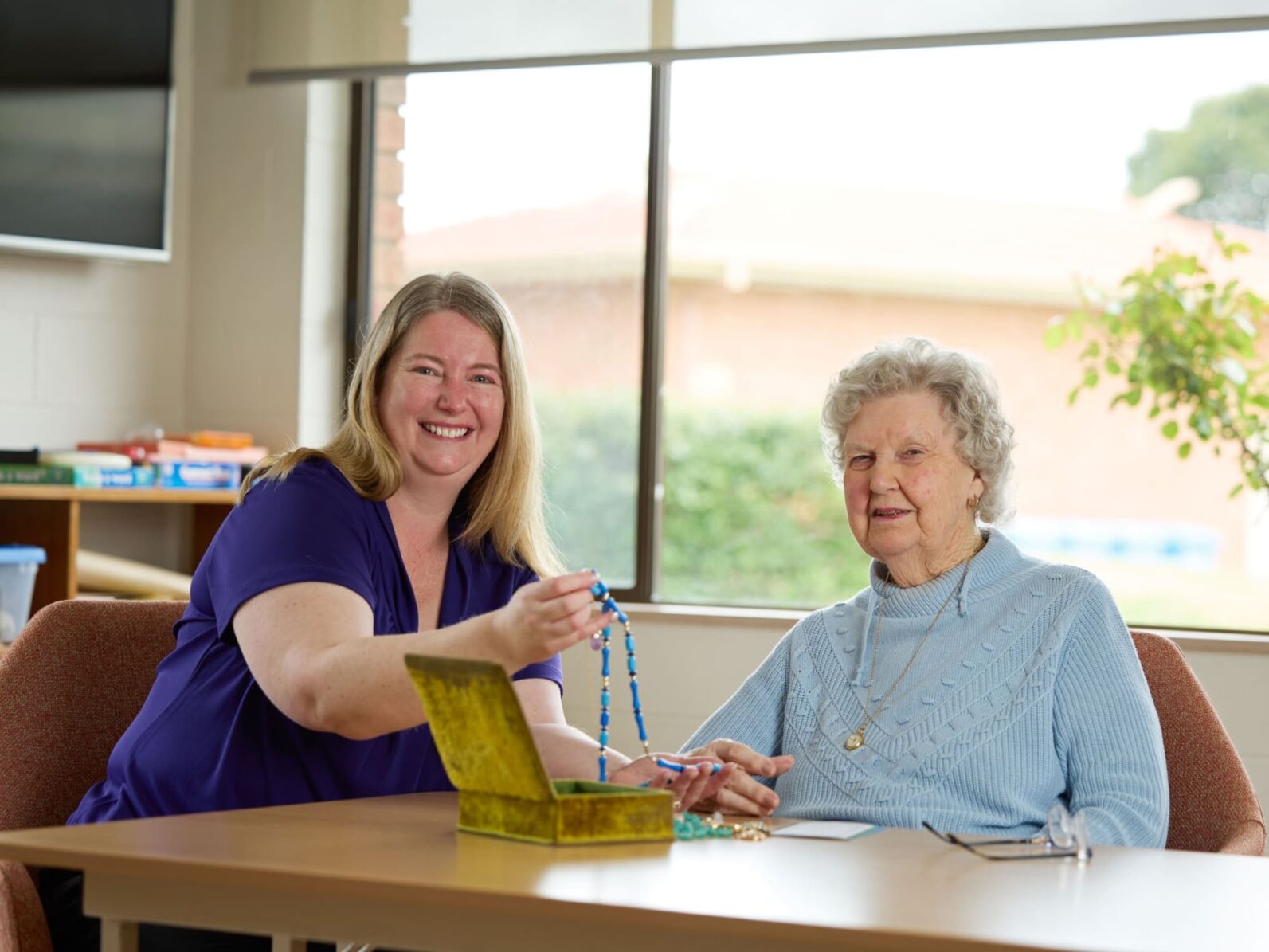 A Uniting AgeWell staff member and a resident sitting at a table, smiling at the camera.