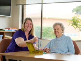 A Uniting AgeWell staff member and a resident sitting at a table, smiling at the camera.
