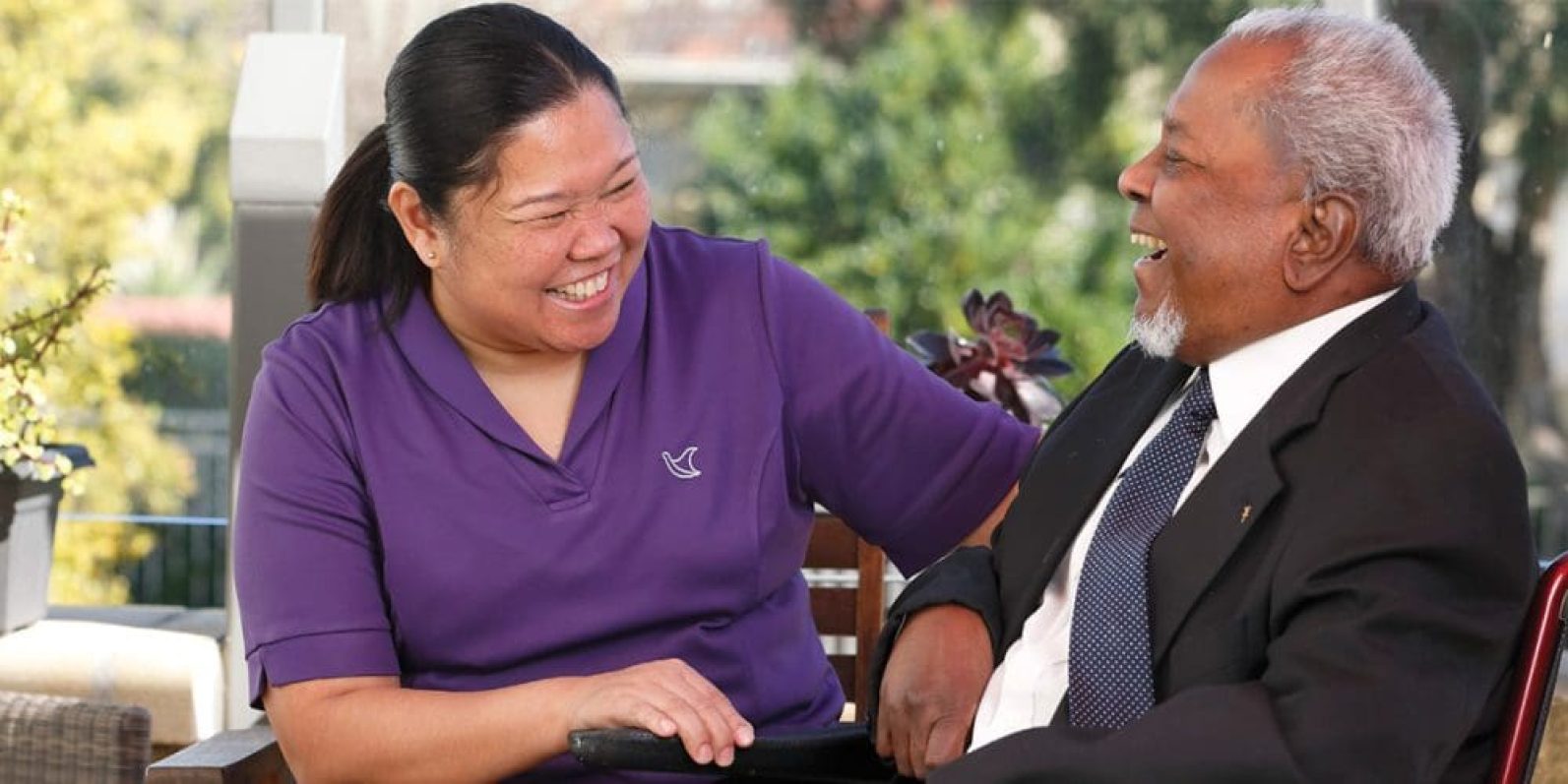 A staff member wearing a purple uniform top sits beside an older adult in a suit jacket and tie. Both are seated indoors near large windows overlooking greenery, engaged in conversation in a bright and welcoming setting that highlights companionship, support and social connection.