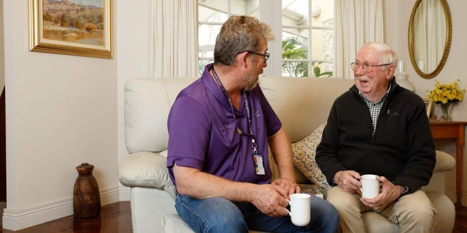 A staff member wearing a purple polo shirt sits on a lounge beside an older adult in a comfortable living room. Both hold mugs and are engaged in conversation, with armchairs, cushions, framed artwork and a large window creating a warm and welcoming home environment.