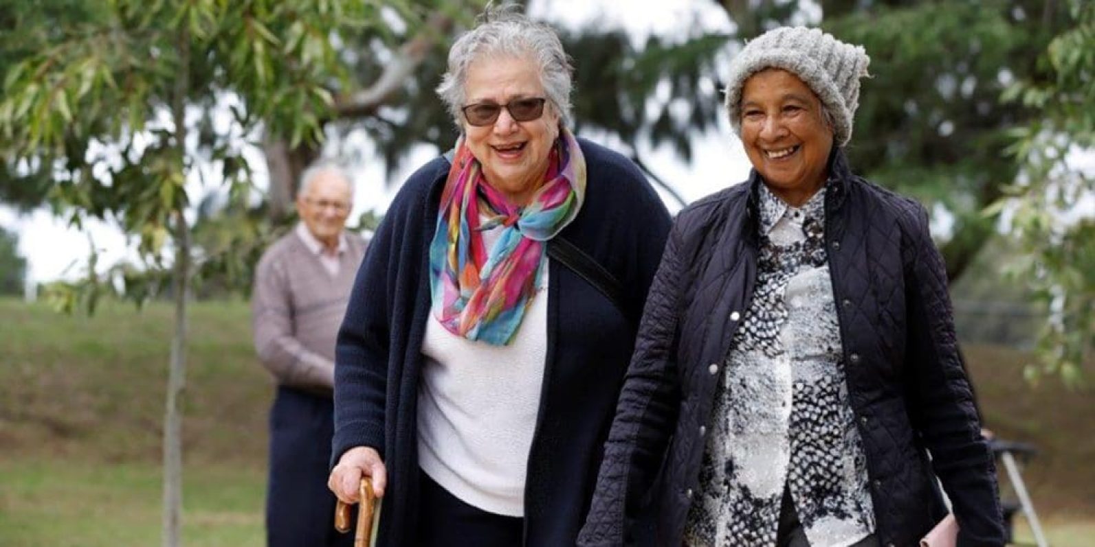 Two people walk side by side through a leafy outdoor garden area. One person uses a walking stick and wears a colourful scarf, while the other wears a knitted beanie and quilted jacket. Another person is visible in the background among the trees and greenery, creating a relaxed and social outdoor setting.