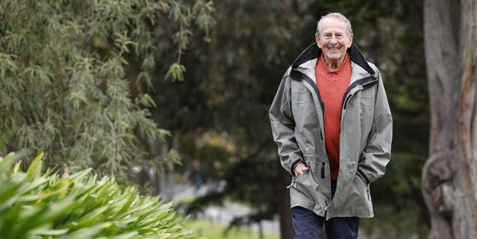 A person wearing a grey jacket over a red top walks along a garden path surrounded by trees and greenery. The outdoor setting features mature plants and natural landscaping, creating a peaceful environment for walking and enjoying the outdoors.