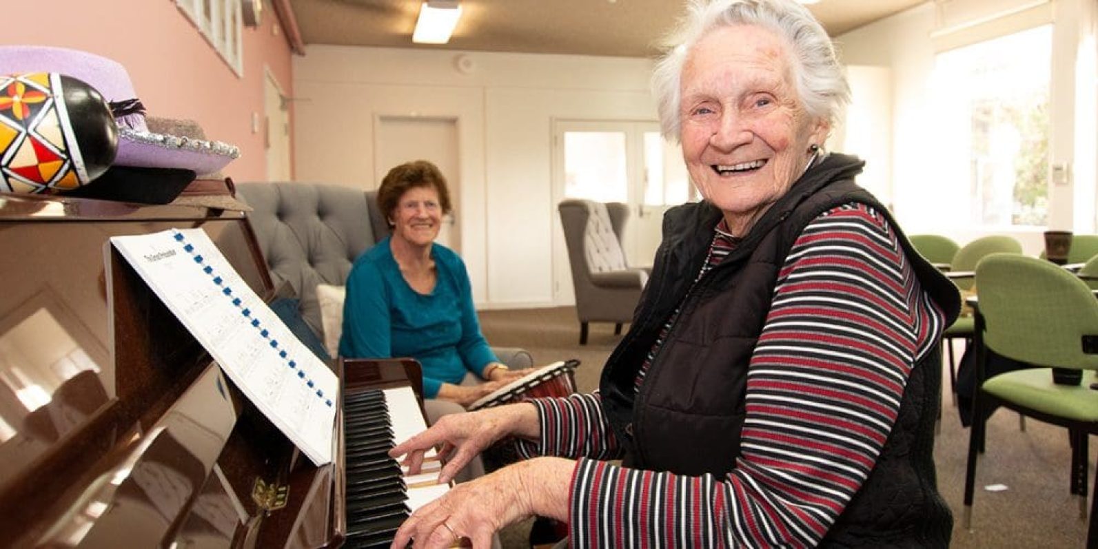 A person sits at a piano in a communal lounge, playing music while another person sits nearby holding a small drum. Comfortable seating, natural light and activity tables create a welcoming environment for music and social engagement.