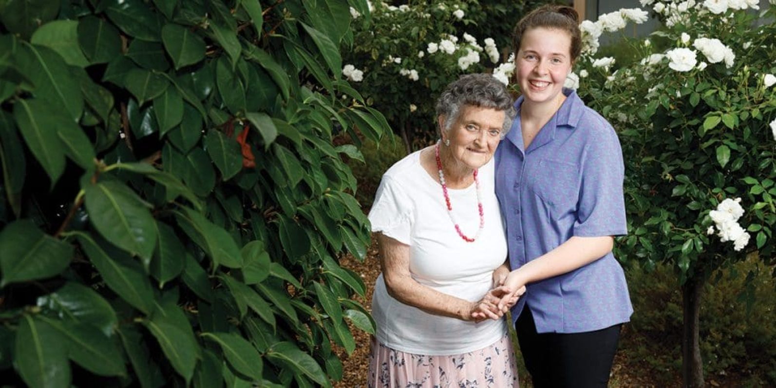 An older adult and a staff member stand together in a garden pathway surrounded by lush green foliage and blooming white roses. The pair hold hands and pose among the landscaped garden beds, highlighting companionship, support and connection in an outdoor setting.