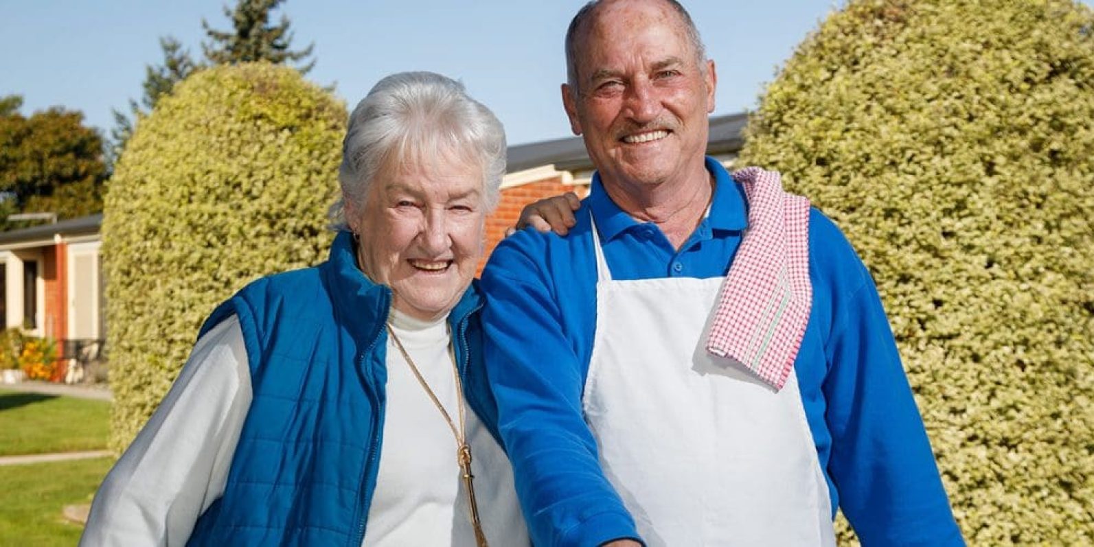 Two people stand together outdoors in a landscaped courtyard with lawns, shrubs and residential-style buildings in the background. One person wears a blue vest and the other a white apron over a blue shirt, with a tea towel draped over one shoulder. The pair are positioned side by side in a sunny garden setting.