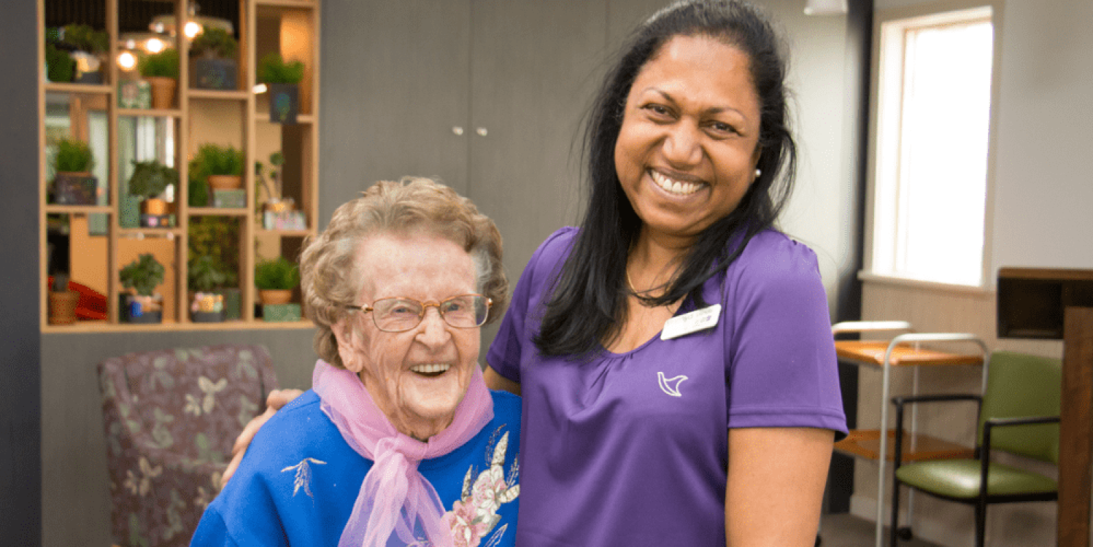 A staff member wearing a purple uniform top stands beside an older adult wearing glasses, a blue floral top and a pink scarf. The pair are standing together in a bright communal lounge, with decorative shelving, indoor plants and seating visible in the background.