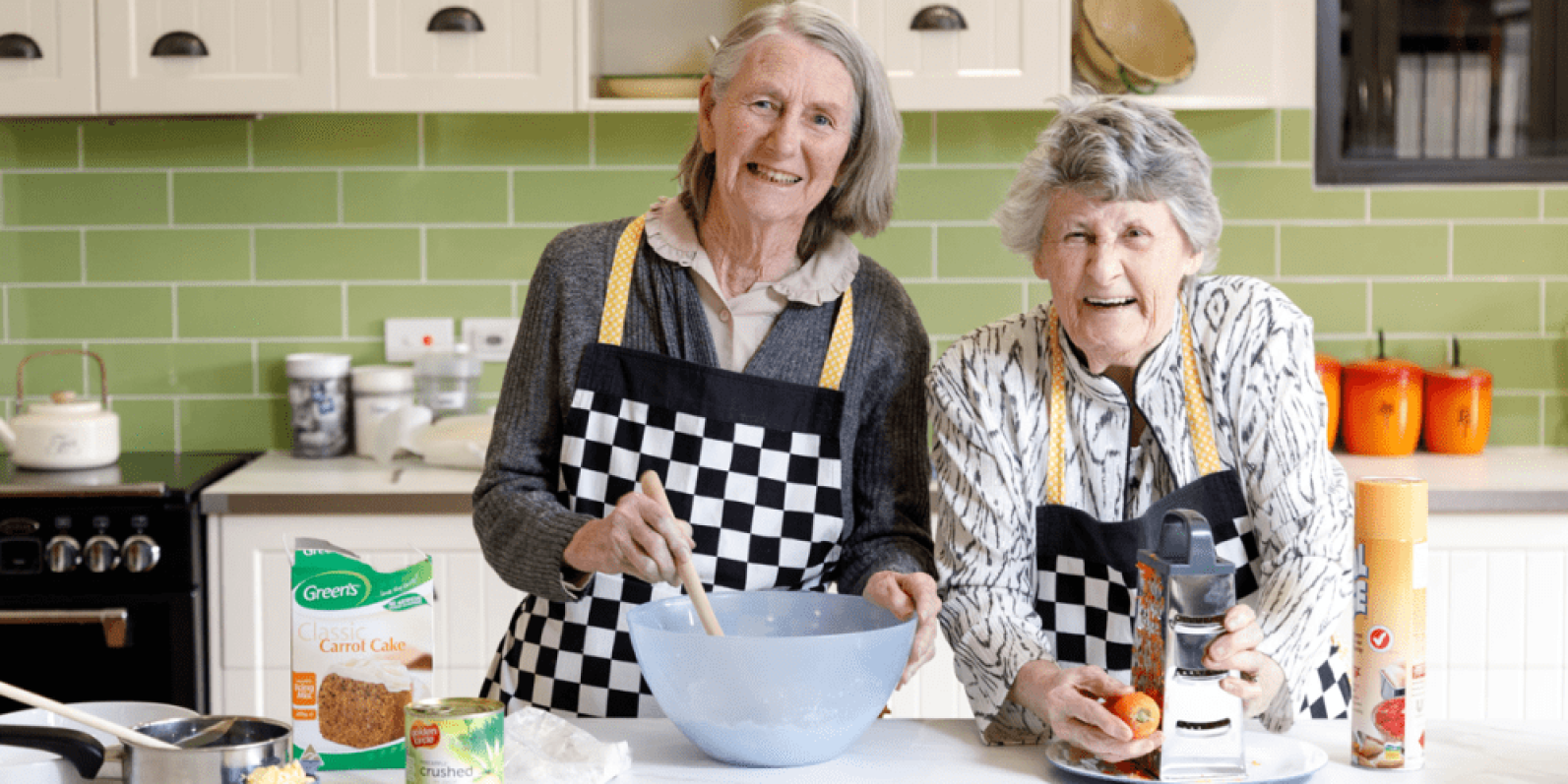 Two older adults wearing checkered aprons prepare food together in a bright kitchen. One person stirs ingredients in a mixing bowl while the other uses a grater, with baking ingredients and cooking utensils arranged on the benchtop. Green tiled splashbacks and cream cabinetry create a warm and homely setting.