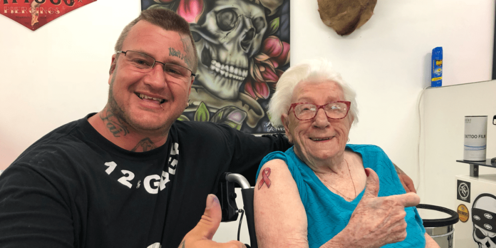A person and an older adult sit together in a tattoo studio, both giving a thumbs-up gesture. The older adult is seated in a wheelchair and displays a small pink ribbon tattoo on their upper arm. Tattoo artwork, studio equipment and decorative wall pieces are visible in the background.