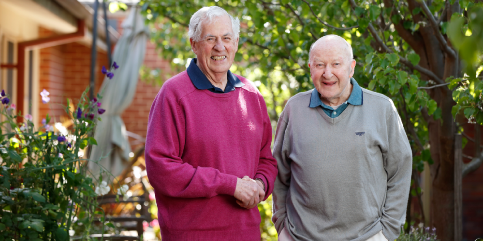 Two older adults stand together in a leafy garden courtyard surrounded by trees, flowering plants and outdoor seating. One person wears a bright pink jumper and the other a light grey jumper, with both positioned on a pathway in a welcoming outdoor setting.