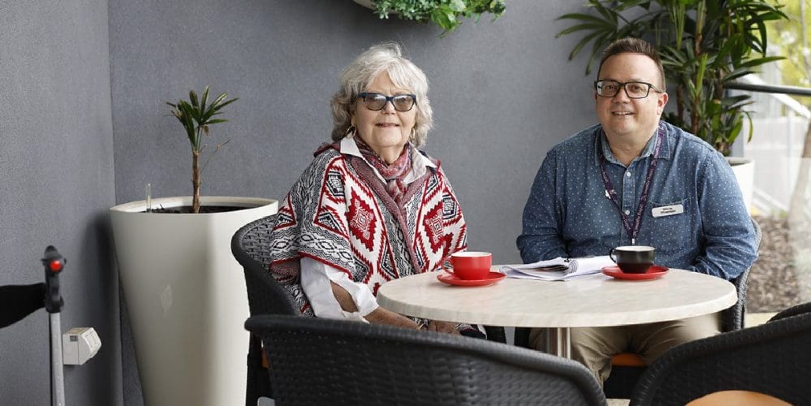 A person wearing glasses and a patterned poncho sits at an outdoor café-style table with a staff member. Two cups sit on the table alongside paperwork, while potted plants and a covered courtyard setting create a welcoming space for conversation and social connection.